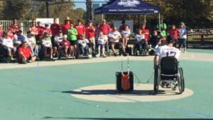 A coach sitting in a wheel chair beside a speaker addressing disables sports members in an open court